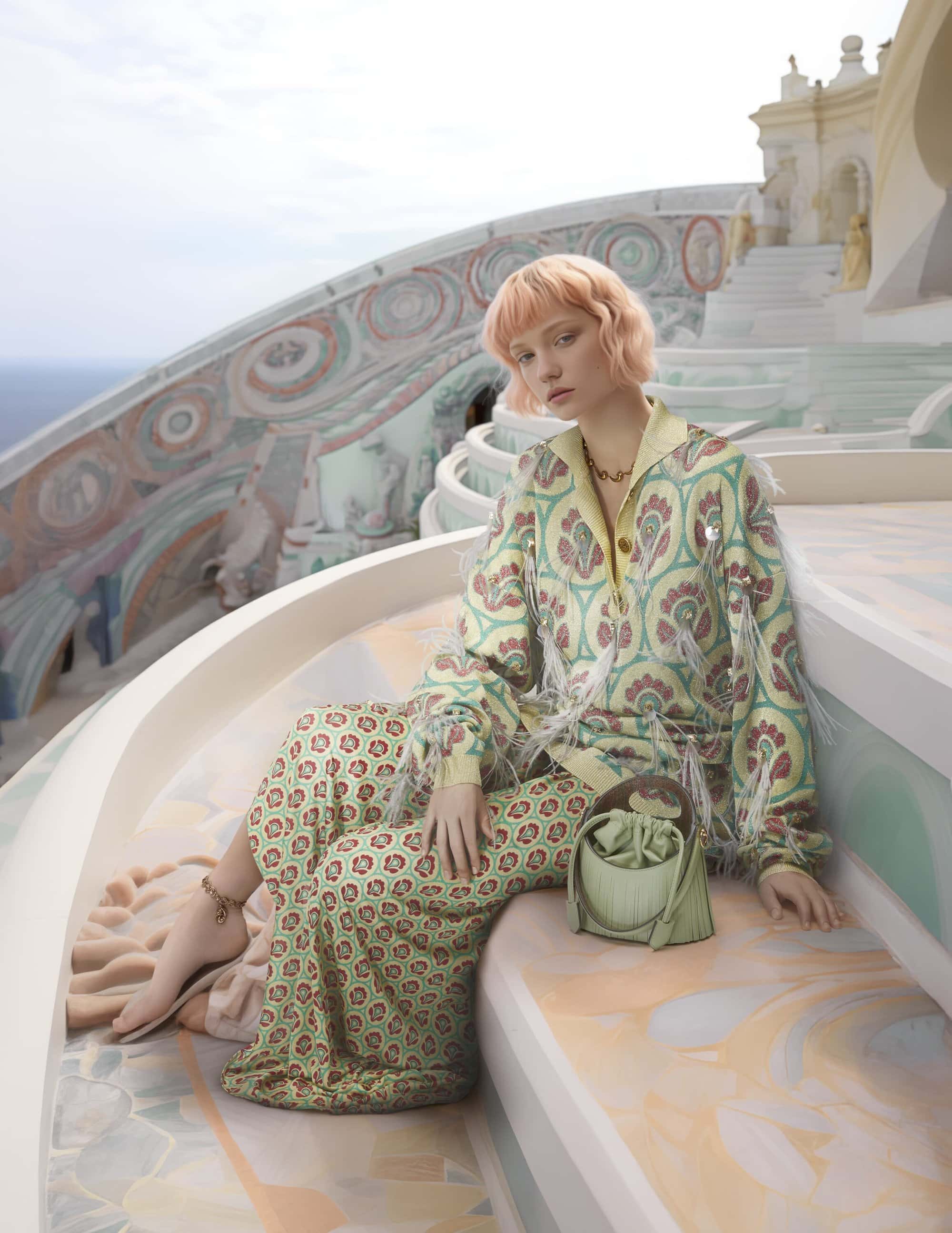 A woman in a printed jacket and pants poses on patterned steps next to a light green purse in an outdoor daytime fashion shot.