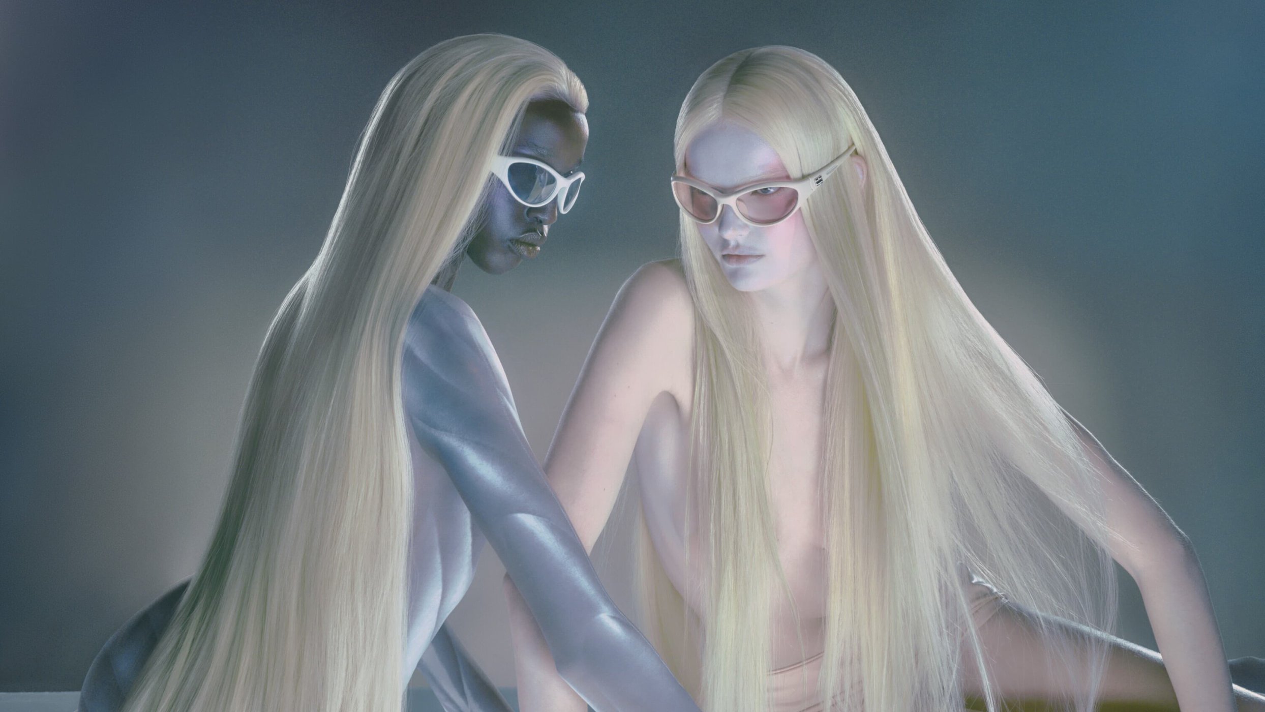 Two fashion models with fair skin and long blond hair posing in sunglasses in a photo studio against a grey-blue background. Beauty shot.