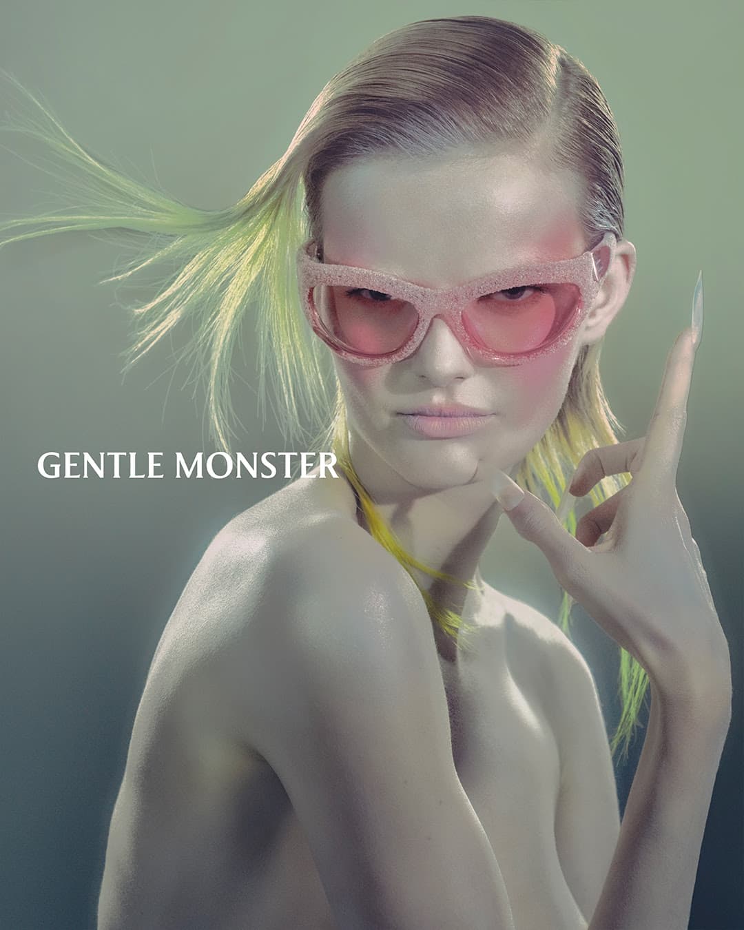Close-up of a woman with pink sunglasses and pale skin, posing with long fingernails, against a muted green studio background. Eyewear fashion shot.