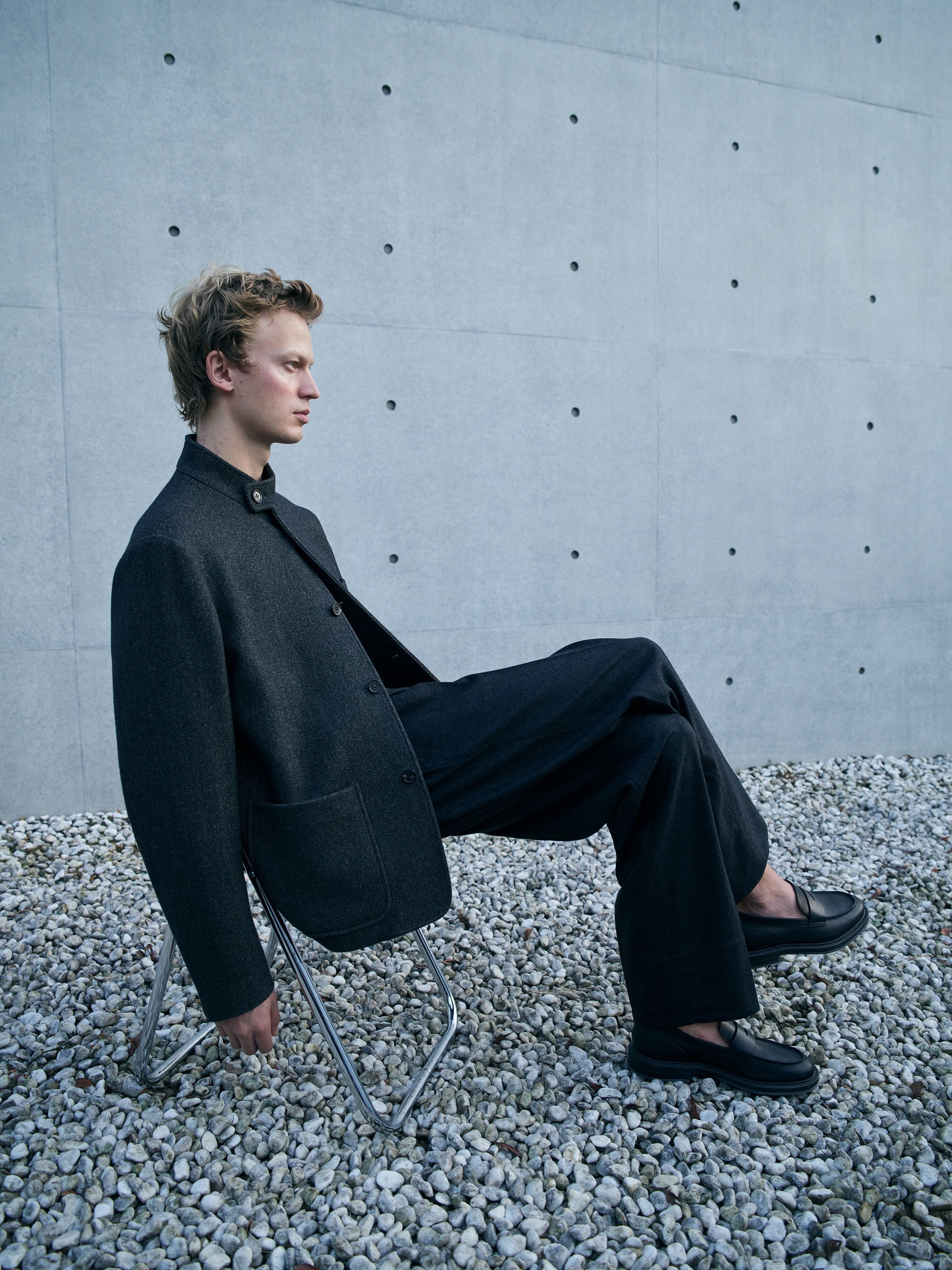 A young man with blond hair sits on a folding chair outdoors, wearing a dark blazer, wide-legged pants, and loafers against a grey wall.