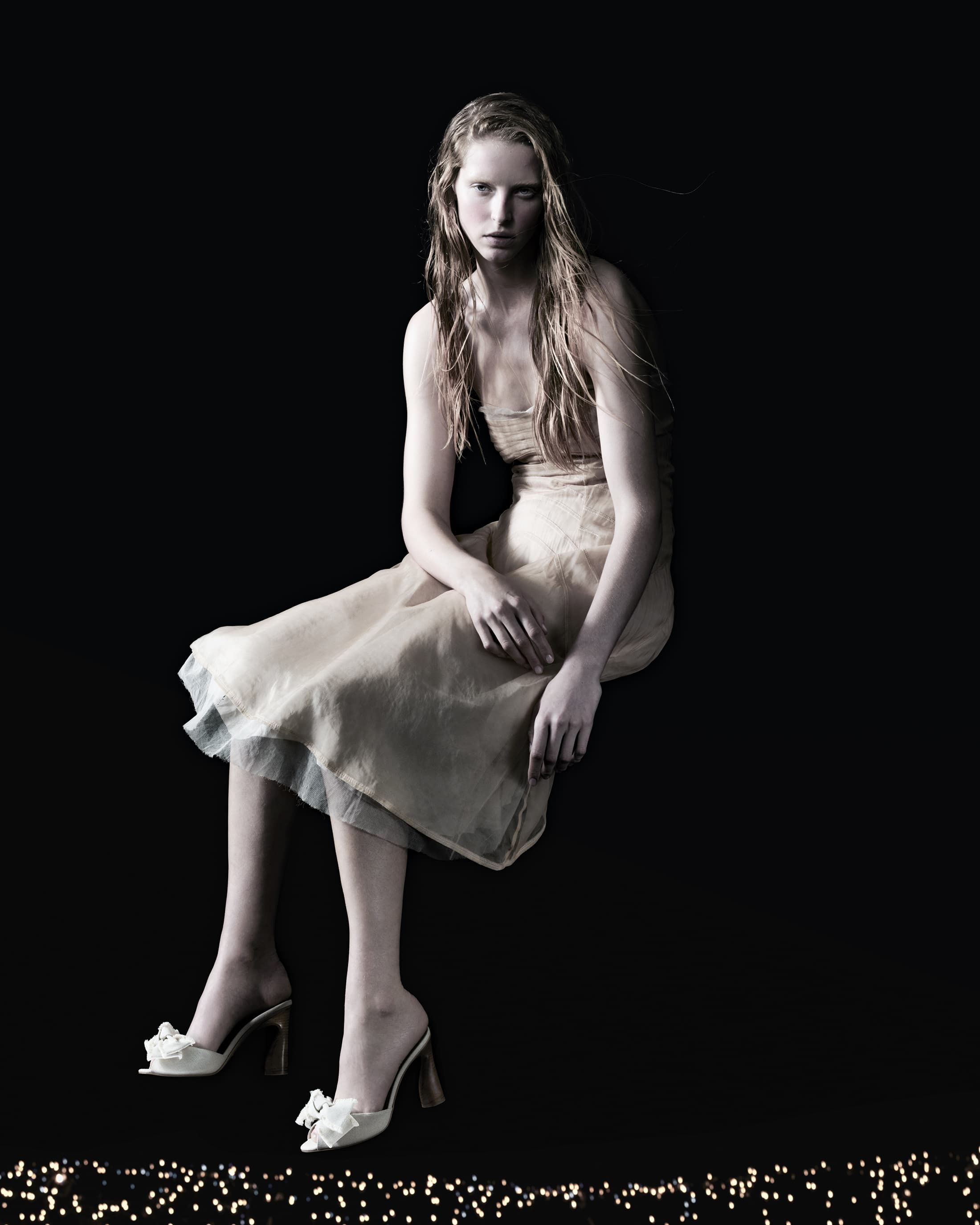 A blonde woman in a dress sits against a dark background, wearing white shoes with bows, accented by a string of small lights.