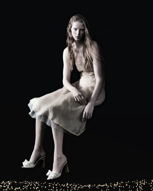 A blonde woman in a dress sits against a dark background, wearing white shoes with bows, accented by a string of small lights.