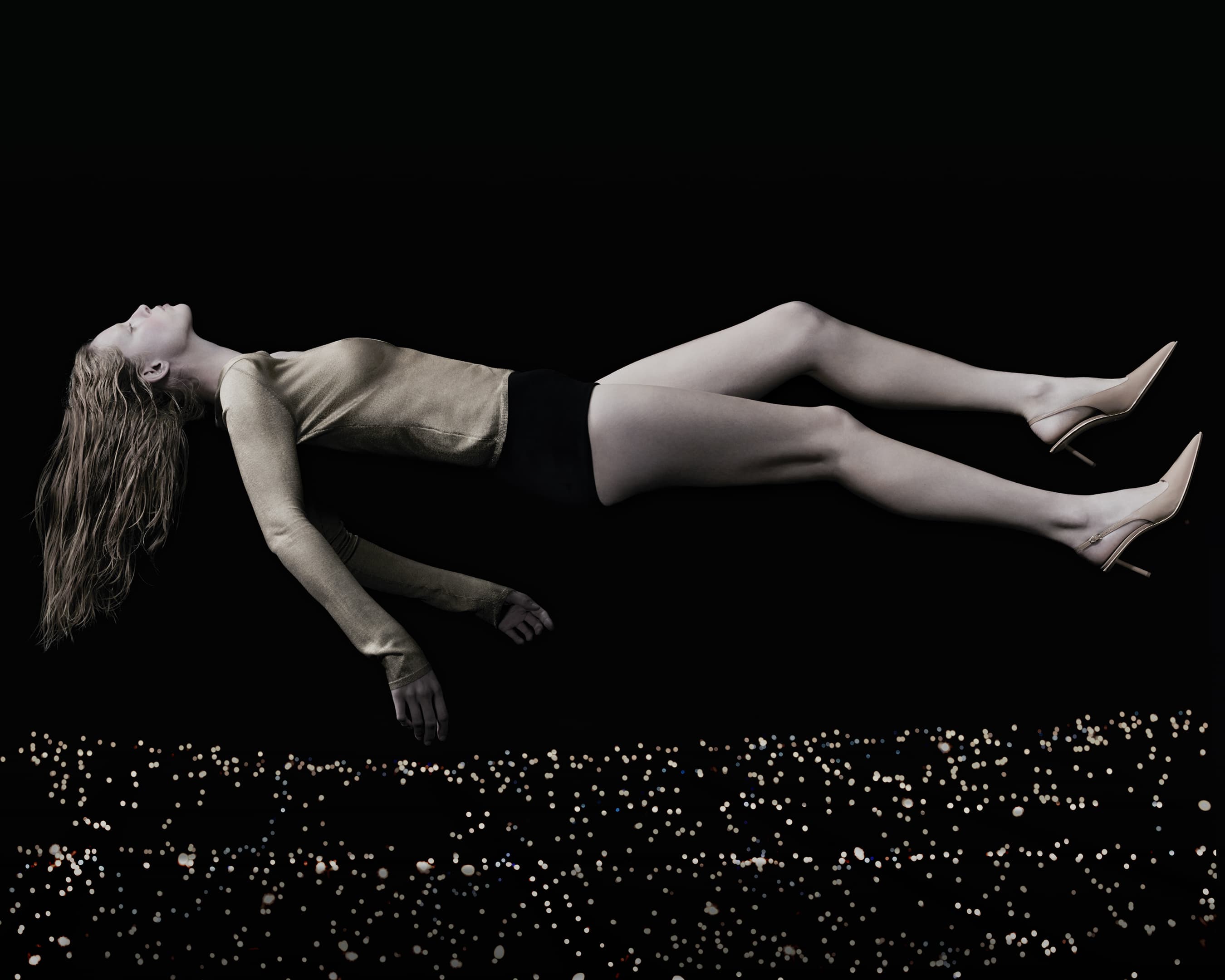 A woman in a gold long-sleeved shirt and black shorts is floating with her blonde hair down over a dark background with light effects.