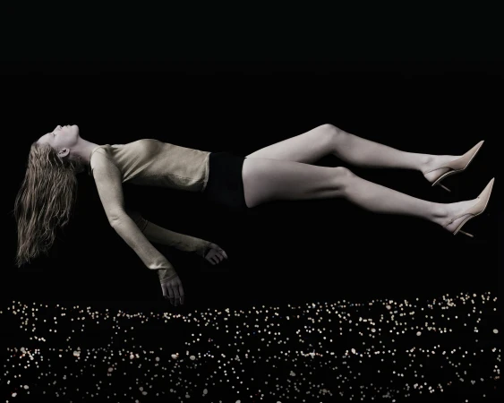A woman in a gold long-sleeved shirt and black shorts is floating with her blonde hair down over a dark background with light effects.