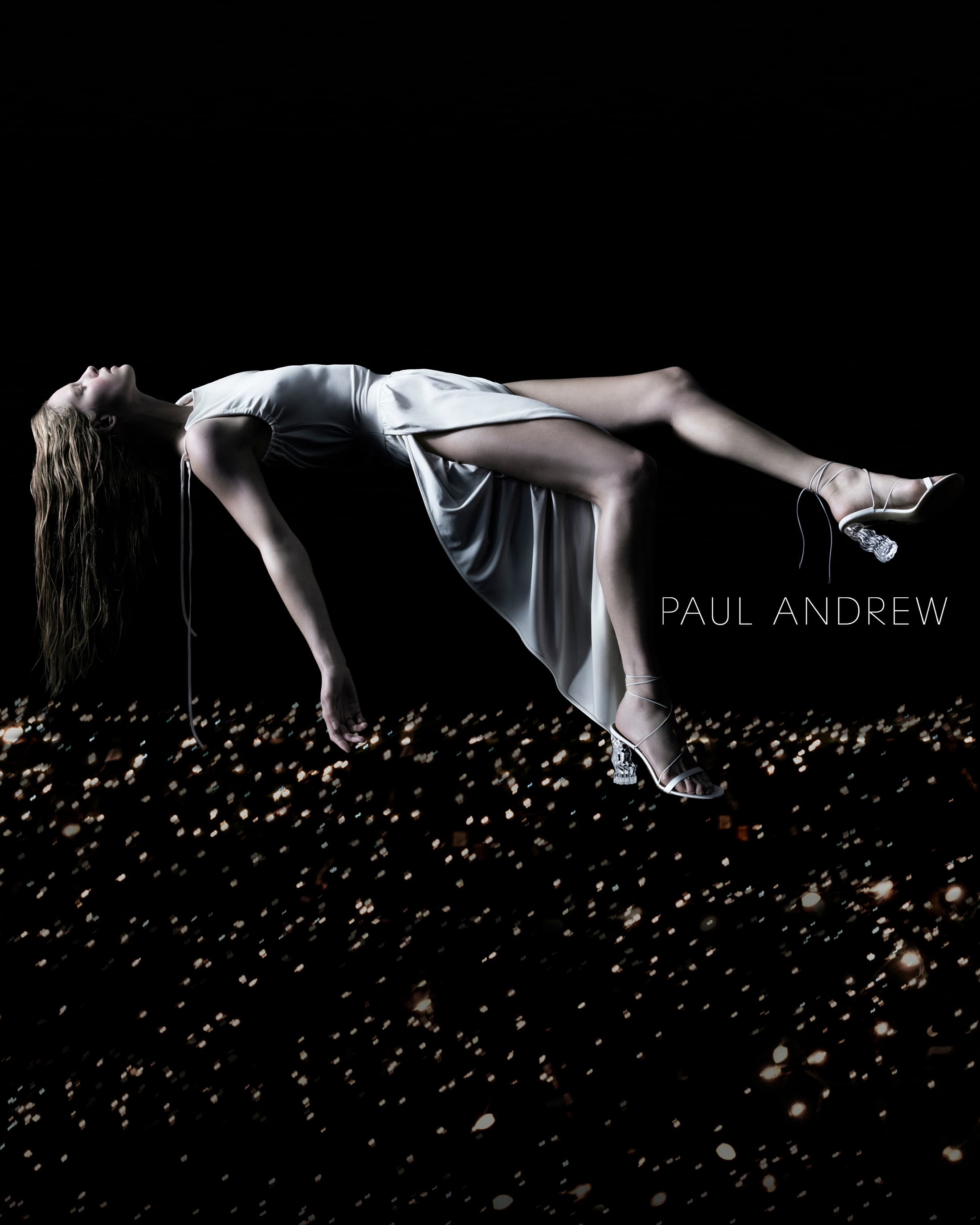 A woman in a dress and heels is suspended in the air over a field of lights against a black backdrop in a studio fashion shot.