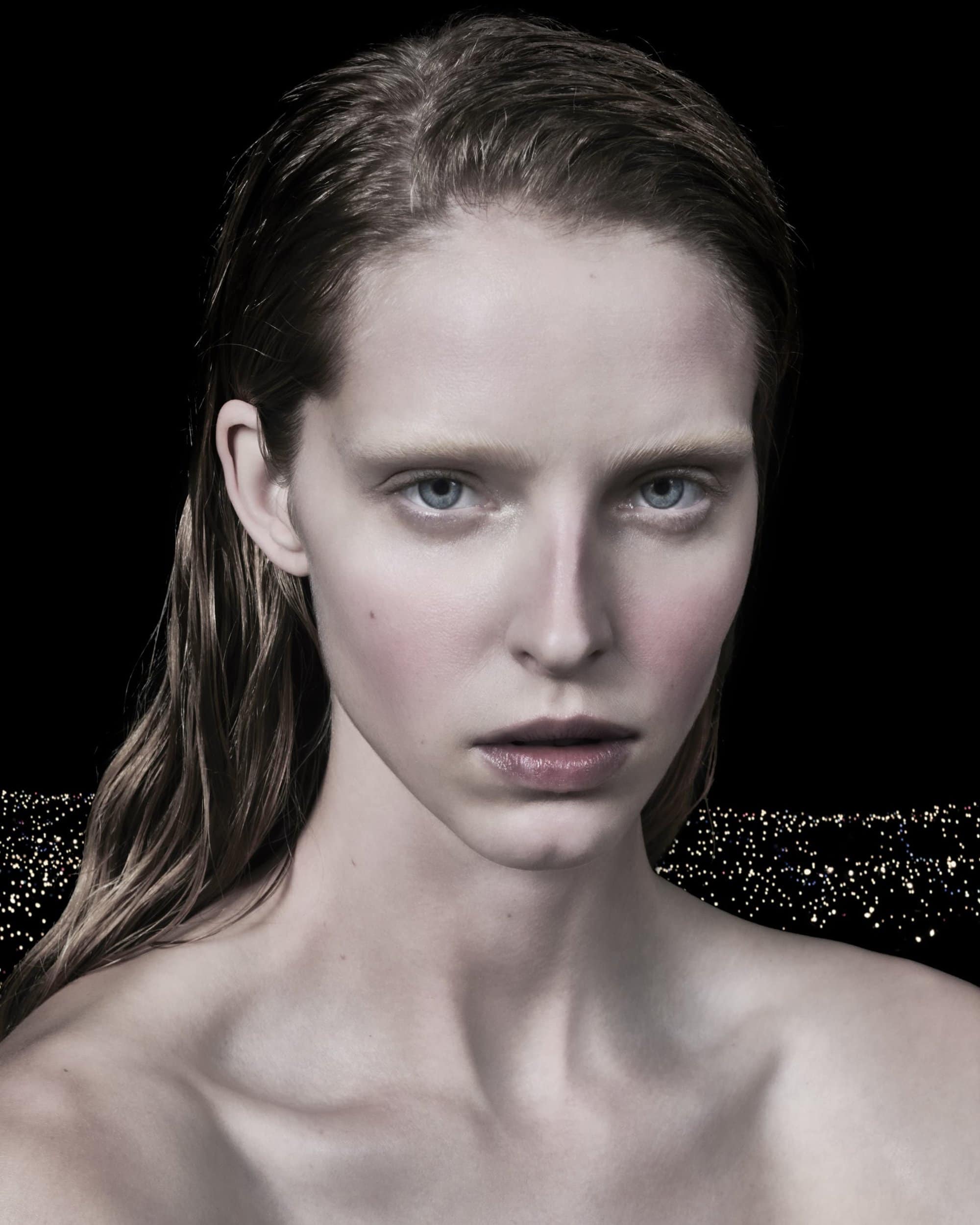 Close-up studio portrait of a young adult woman with light skin and wet hair against a dark background with abstract lights.
