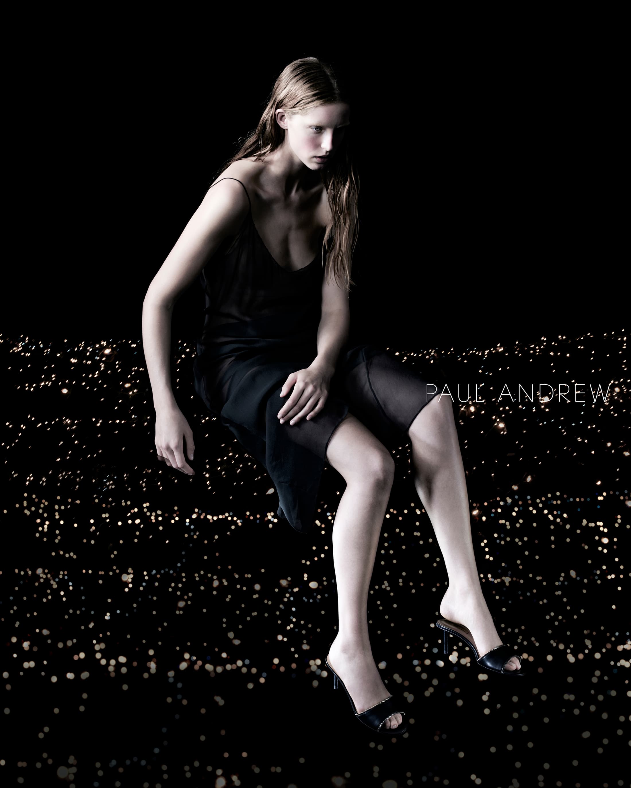 A woman wearing a dress and heels sits against a dark background with a scattering of small lights, creating a moody fashion shot.