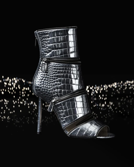 High-heeled peep-toe ankle bootie in silver with crocodile texture and zipper details, shot against a dark background with bokeh effect.
