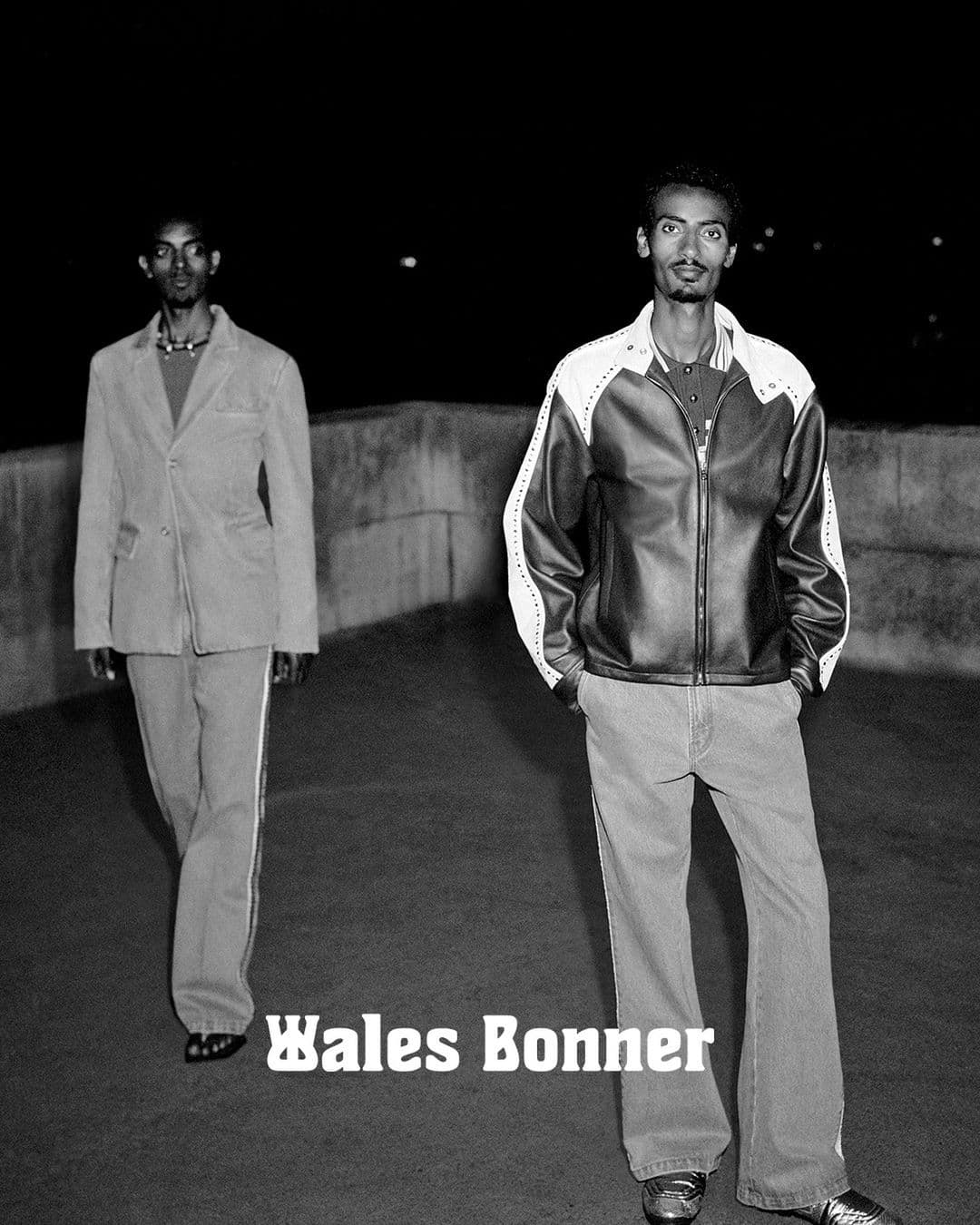 Two men stand outdoors in Wales Bonner fashion, one in a blazer suit and the other in a leather jacket and jeans, beneath a dark sky.