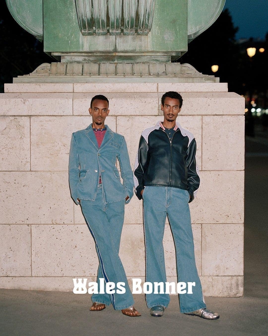 Two adult men standing side-by-side in denim and leather jackets in a city setting, showcasing fashion and urban style at night.
