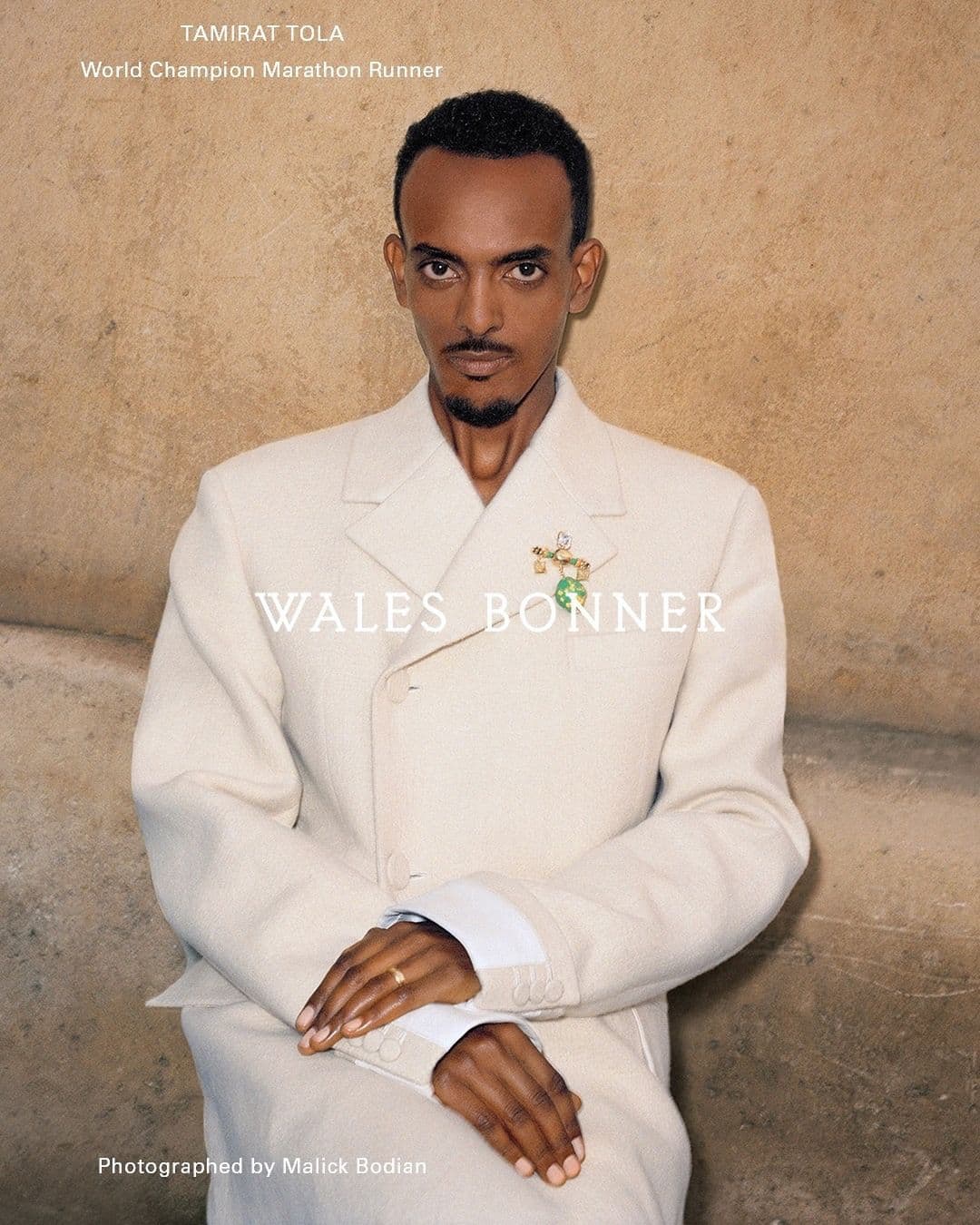 Portrait of Tamirat Tola, world champion marathon runner, in a cream suit against a textured wall backdrop in a studio environment.
