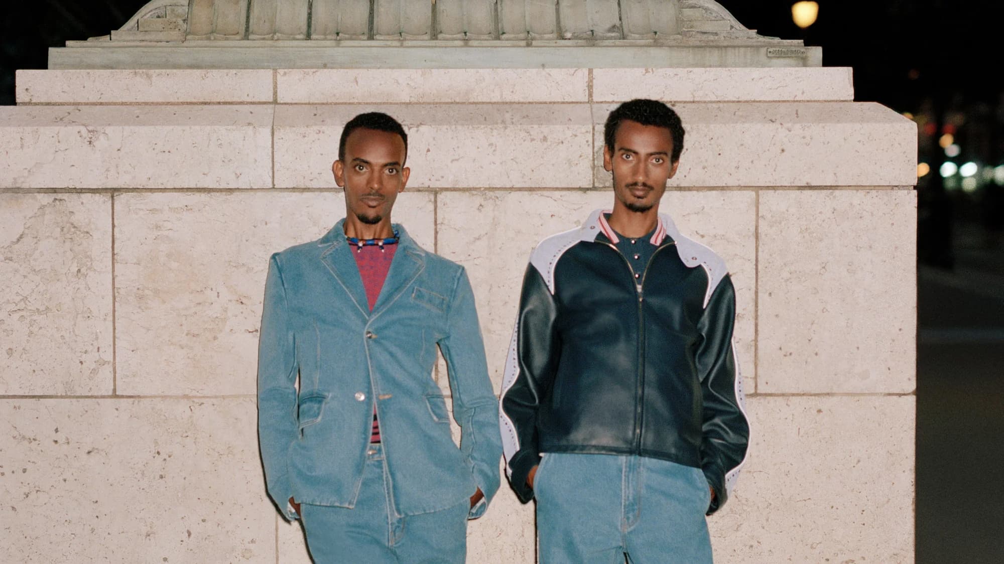 Two dark-skinned men stand outdoors against a stone wall wearing matching light-wash denim jeans and stylish jackets. Cool mood lighting.