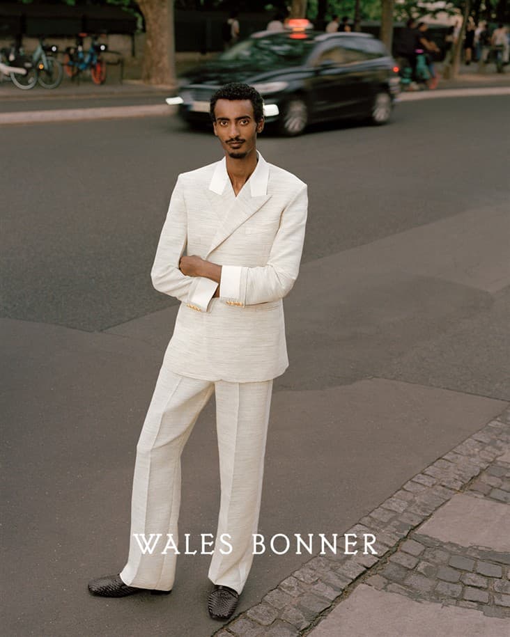 Man in a light-colored suit poses on a city street, standing on the crosswalk with a vehicle and cyclists in the background.