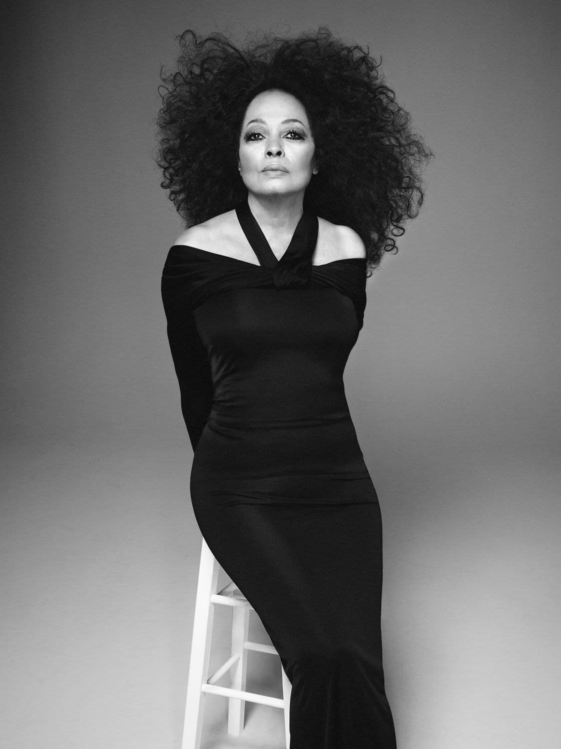 Black and white portrait of a woman with large afro wearing a long black dress, seated on a small white step stool in a studio.