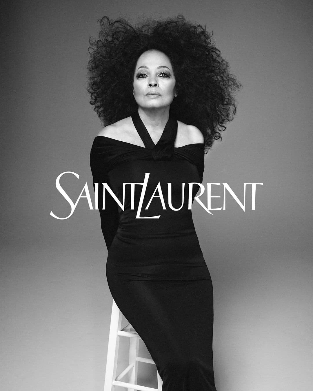 A woman with large curly hair poses in a fitted black dress and sits on a white stool. Saint Laurent logo displayed.