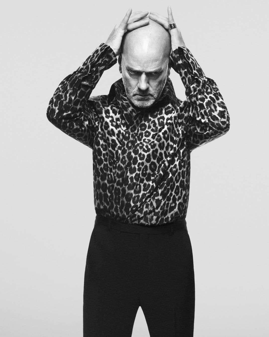 A bald adult male with hands on his head, wearing a leopard print shirt and black pants, poses in front of a grey background.