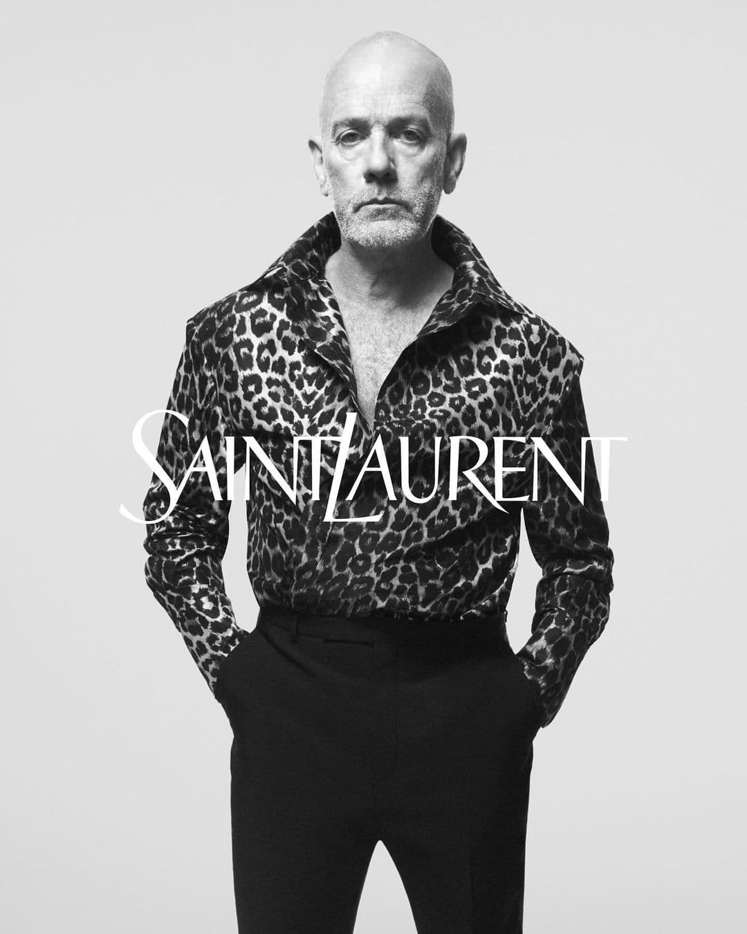 Black and white portrait of a mature man in a leopard print shirt and black pants, hands in pockets, against a plain background.