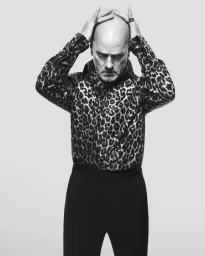 Michael Stipe wearing a leopard-print shirt with hands on his head against a plain grey studio background.