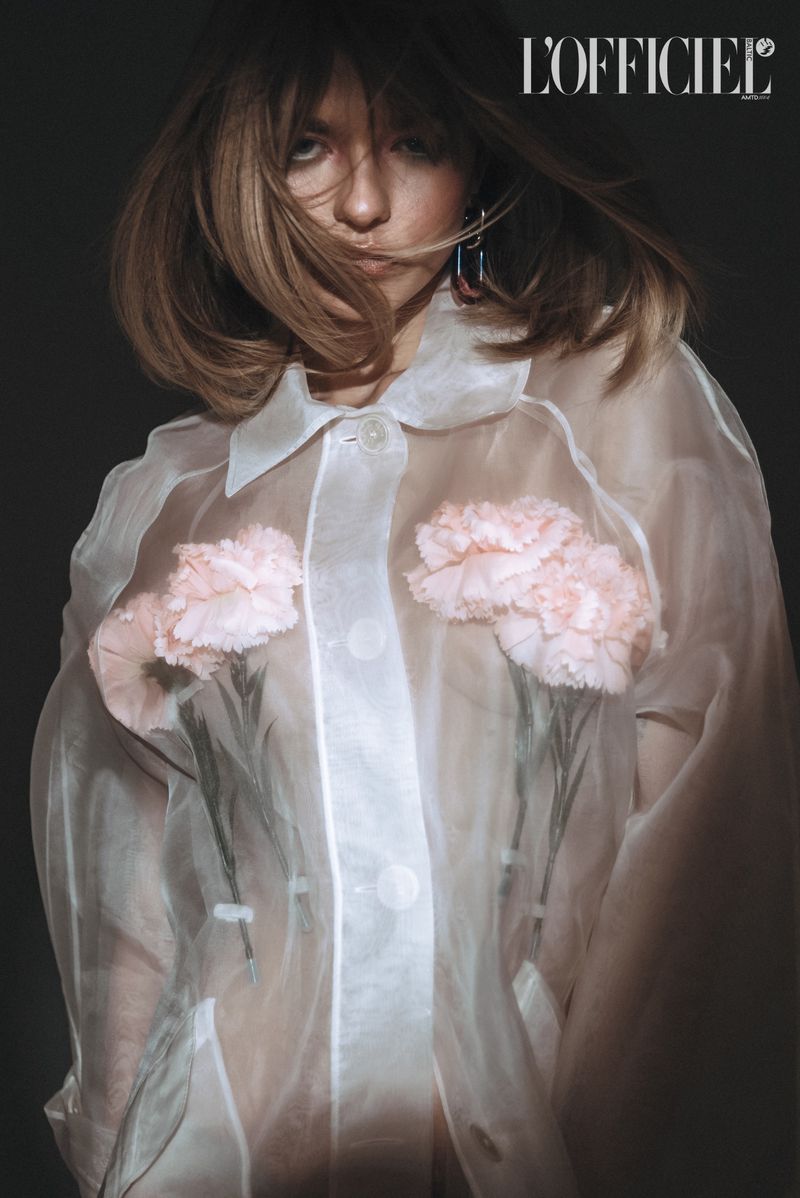 Fashion portrait of an adult female model wearing a sheer white blouse with pink floral details in a studio with a dark backdrop.