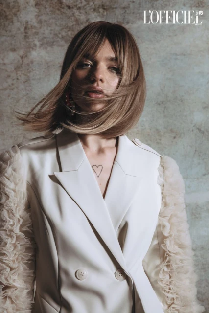 Blonde woman with heart tattoo poses in a white blazer in a beauty and fashion shot against a textured grey background.