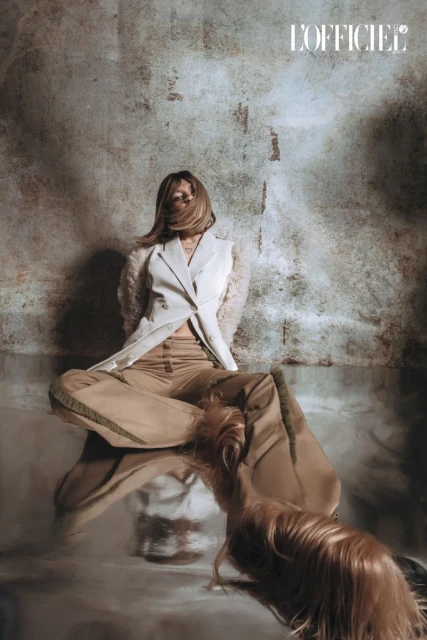 Fashion shot of an adult woman sitting with flowing hair and wearing a white blazer and beige pants in front of a textured wall.