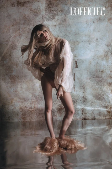 Editorial fashion photo of a slender adult woman posing in a sheer shirt with her feet in hair-covered slippers, posing against a mottled background.