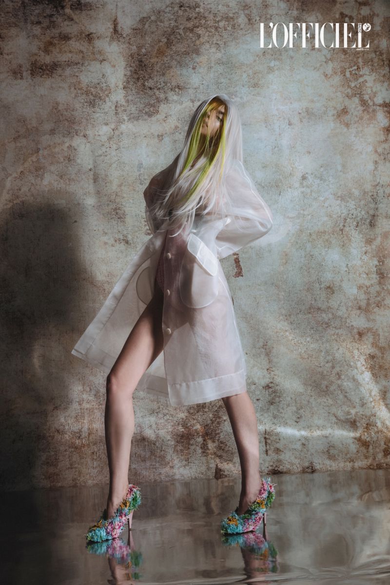 Fashion photography features a model in a transparent coat and floral heels, against a textured background and reflective floor.