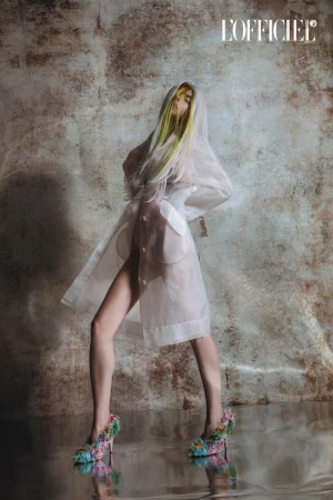 Fashion photography features a model in a transparent coat and floral heels, against a textured background and reflective floor.