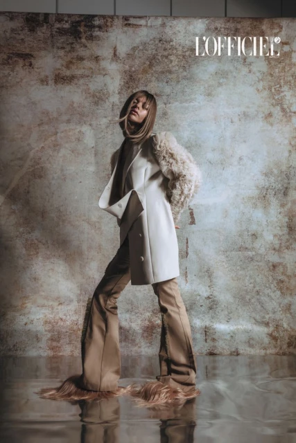 Fashion editorial shot: Woman in white coat and tan pants poses in front of mottled background, magazine logo visible in upper corner.