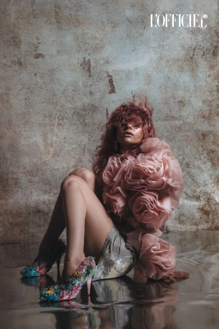 Fashion editorial shot of a seated woman with pink hair and oversized floral top, wearing floral-covered high heels on a reflective surface.