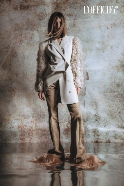 Editorial fashion shot: a woman stands with a neutral expression wearing a white double-breasted blazer, beige pants, and hair on her feet.