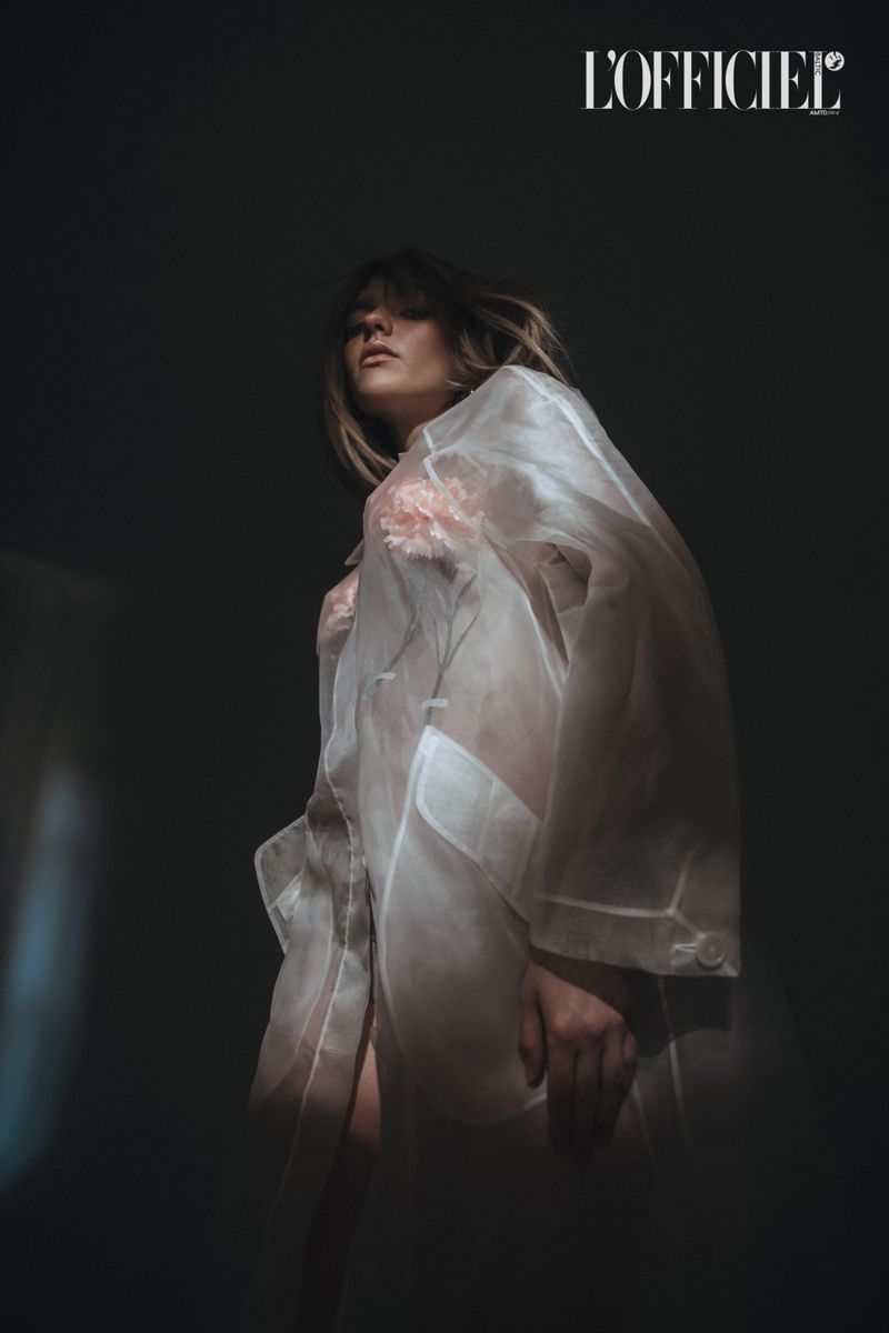 Elegant woman in sheer white robe poses in chiaroscuro light for a fashion shot, with a blurred background and 'L'Officiel' logo visible.