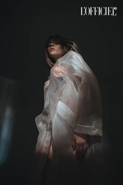 Elegant woman in sheer white robe poses in chiaroscuro light for a fashion shot, with a blurred background and 'L'Officiel' logo visible.