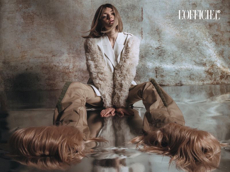 Fashion editorial shot of a fair-skinned woman in a white blazer and khaki pants sitting cross-legged on a reflective surface.