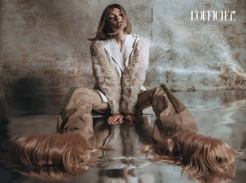 Fashion editorial shot of a fair-skinned woman in a white blazer and khaki pants sitting cross-legged on a reflective surface.