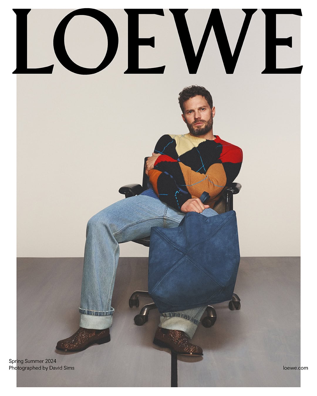 Jamie Dornan sits in a black rolling chair, wearing jeans, a patterned sweater, and patterned boots while holding a blue tote bag.