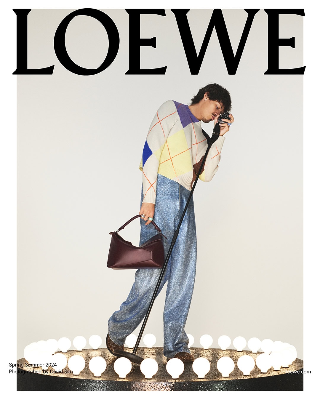 Fashion editorial featuring a man in a patterned sweater, metallic pants, and brown bag, posing under the Loewe logo with bright lights.