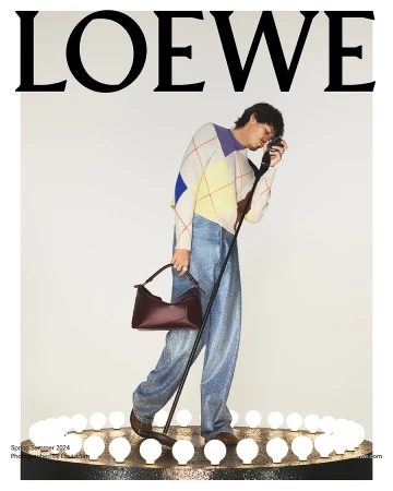 Fashion editorial featuring a man in a patterned sweater, metallic pants, and brown bag, posing under the Loewe logo with bright lights.