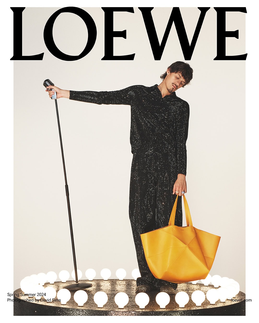 Magazine cover featuring a male model wearing a sparkling black outfit and holding a large yellow tote bag in a studio setting.