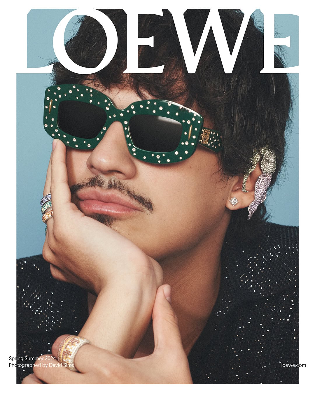 Man with dark curly hair, mustache and dark green glasses with white dots poses in front of a light blue background wearing shiny jewelry.