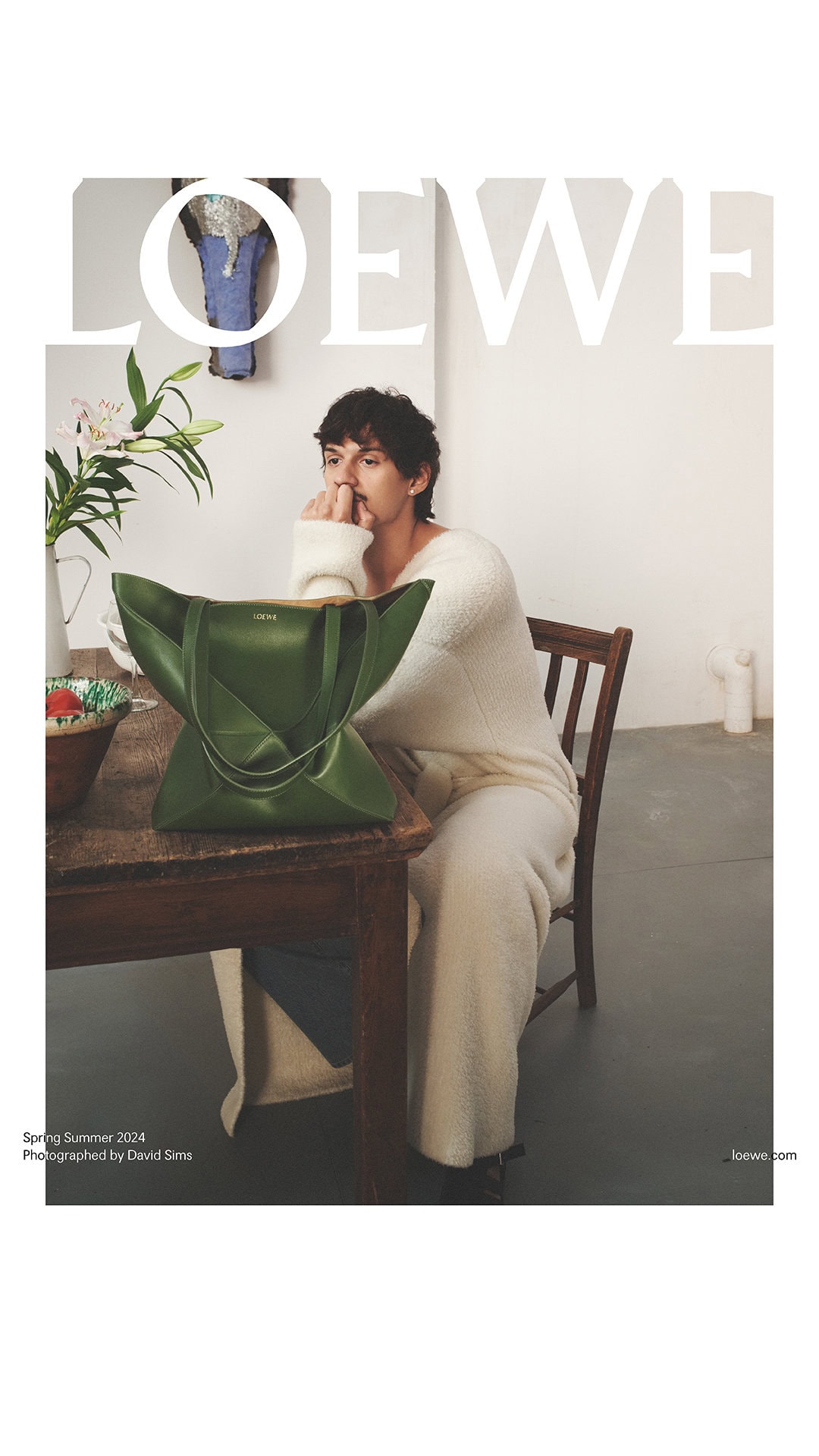 Loewe fashion editorial featuring a male model sitting at a table with a green leather tote bag, lilies, and artwork in a light-filled room.