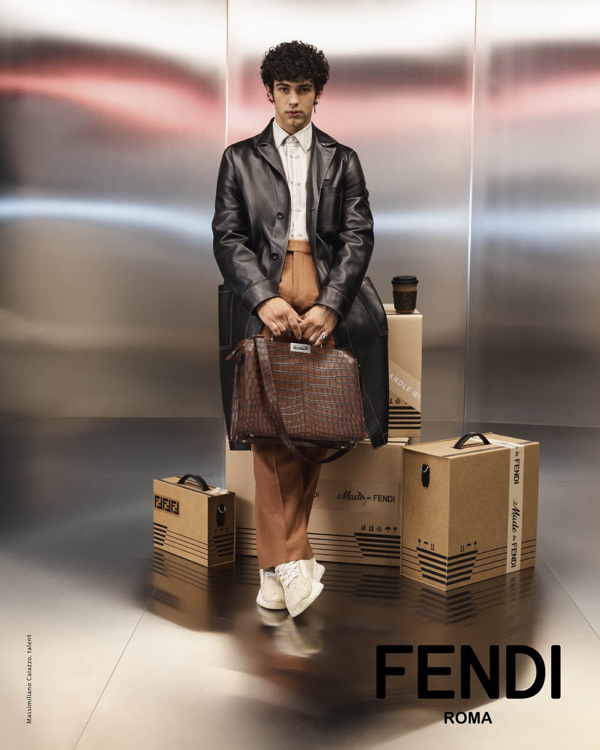 A young adult poses in a leather trench coat, styled with a brown patterned Fendi bag, standing in front of metal panels and boxes.