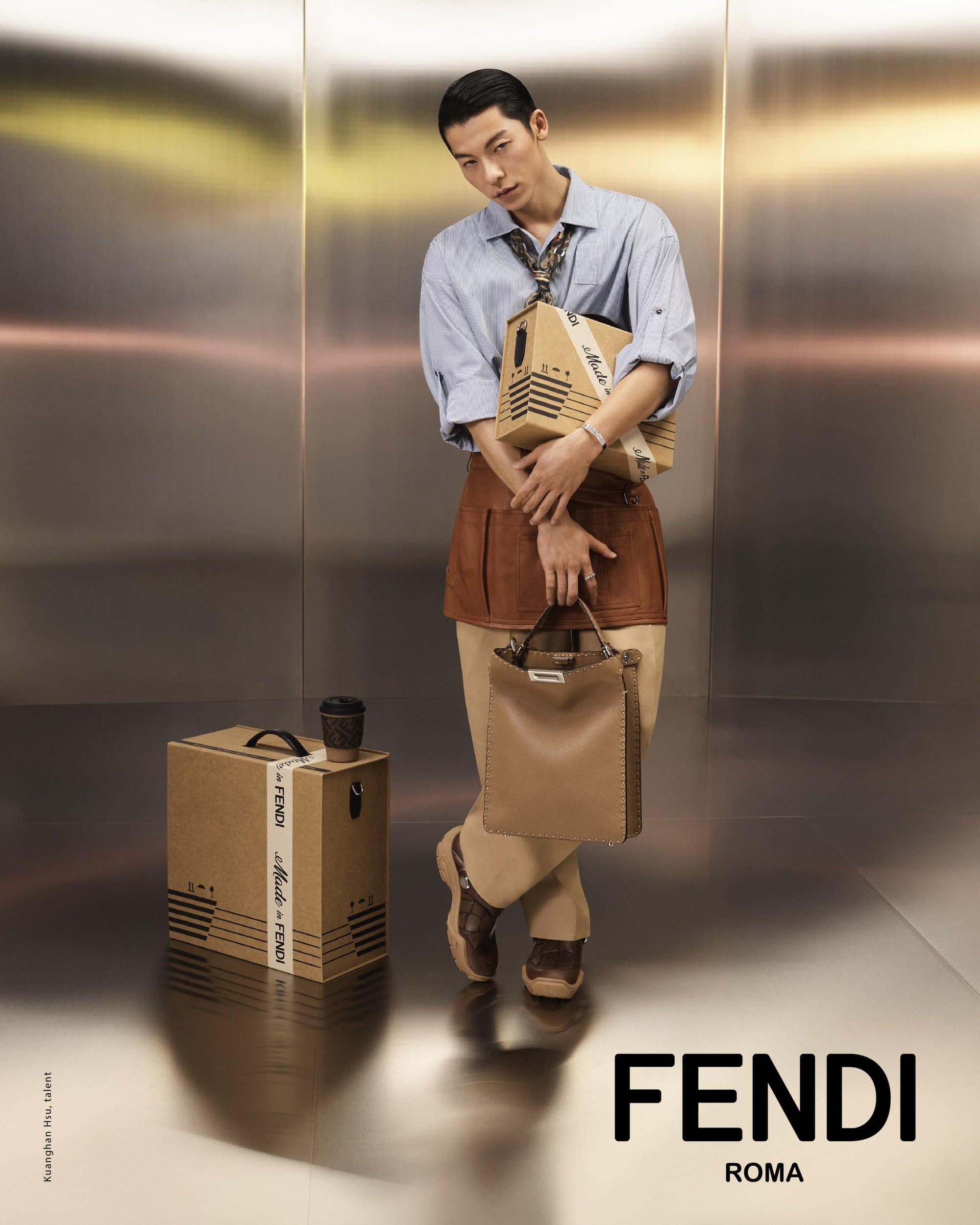 A man with slicked-back hair wears a blue shirt, brown skirt, and loafers, holding a tan Fendi bag and a box. Modern studio setting.