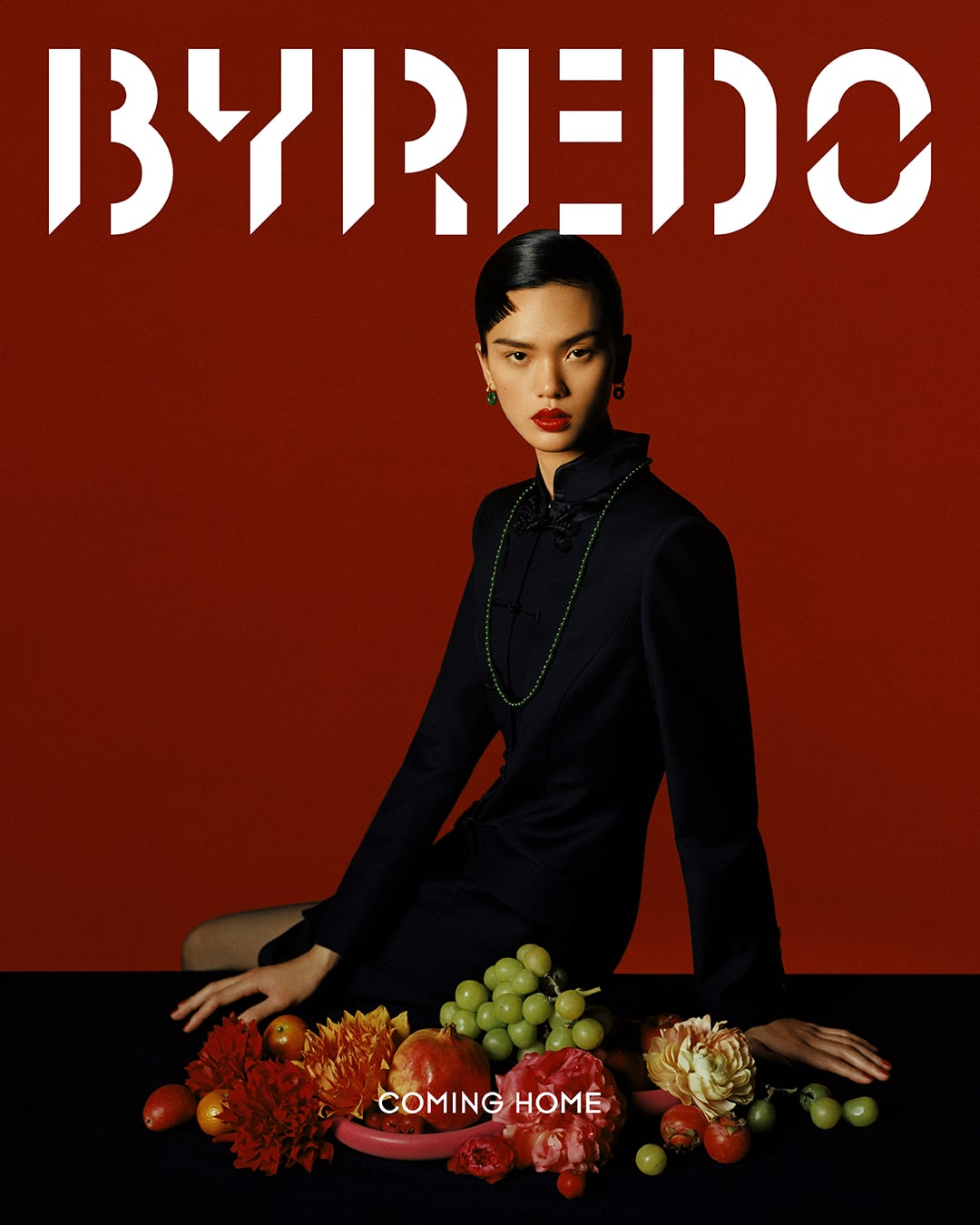 Elegant portrait of a person in a dark dress seated next to a fruit and flower display against a red background, promoting BYREDO.