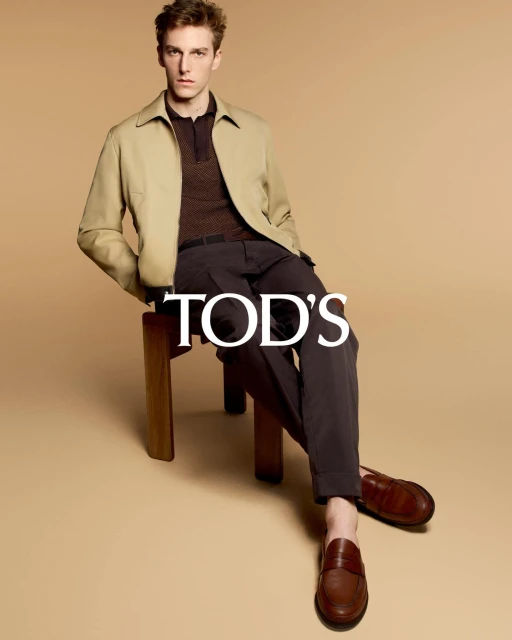A young adult male model sits on a wooden stool wearing a light tan jacket, a dark brown polo shirt, dark slacks, and brown leather loafers.