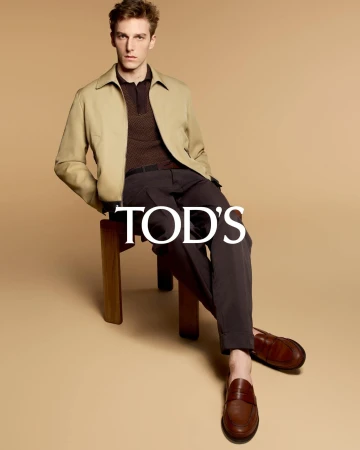 A young adult male model sits on a wooden stool wearing a light tan jacket, a dark brown polo shirt, dark slacks, and brown leather loafers.