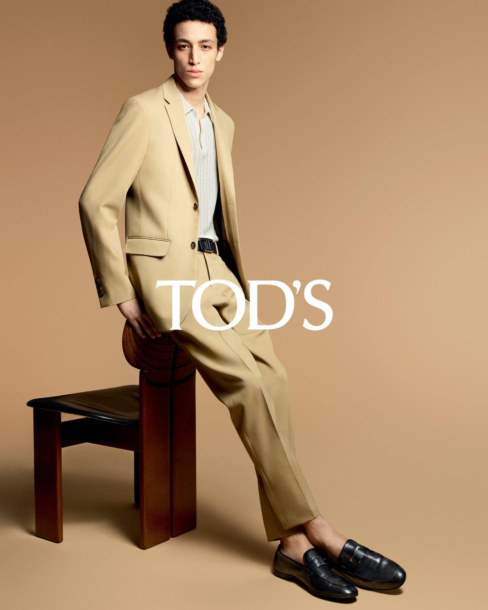 A young adult male in a tan suit poses in a studio setting, seated on a stool. The photograph features a clean, modern aesthetic.