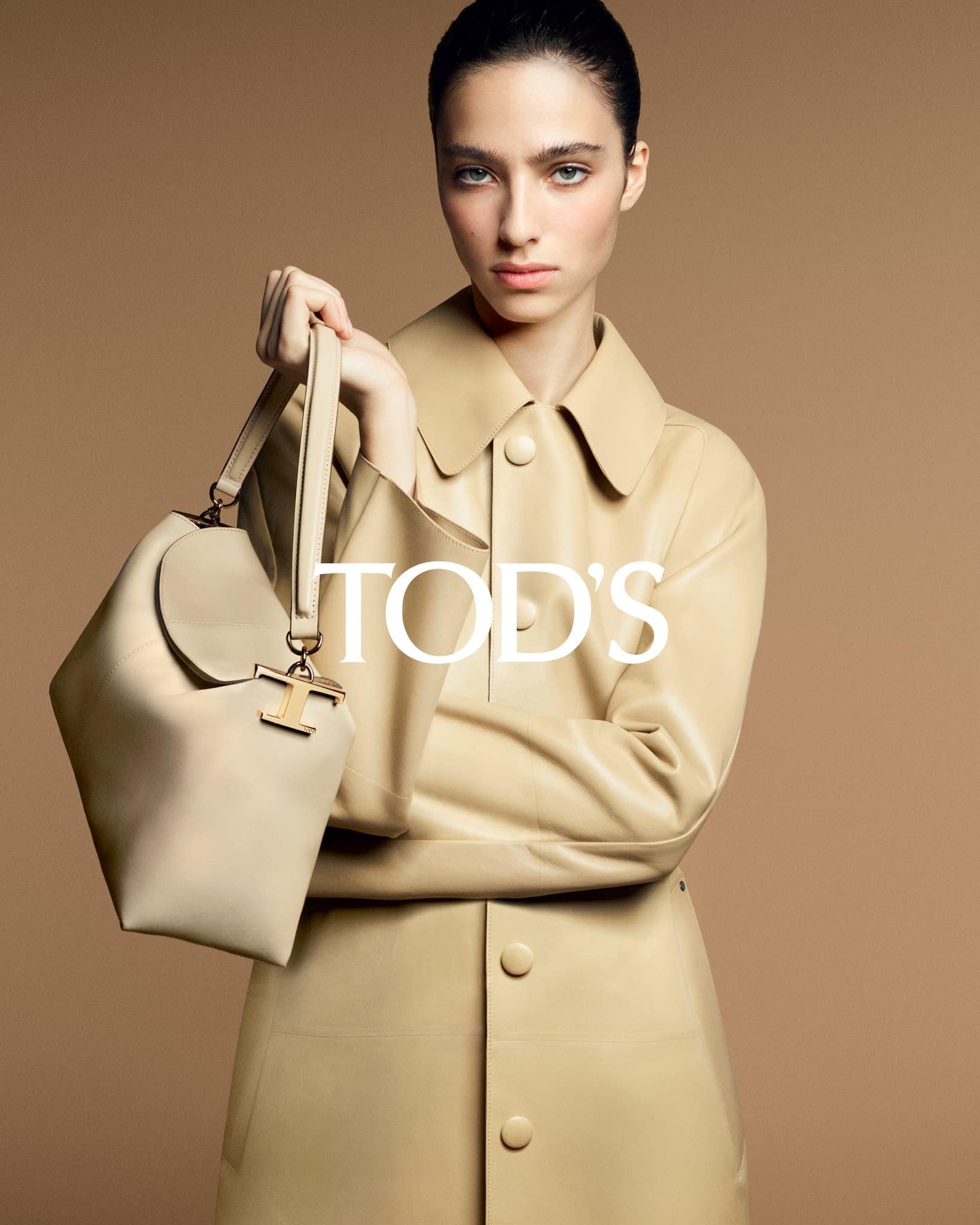 Fashion shot of a woman in a tan coat holding a beige purse with visible logo against a neutral tan background in a studio setting.
