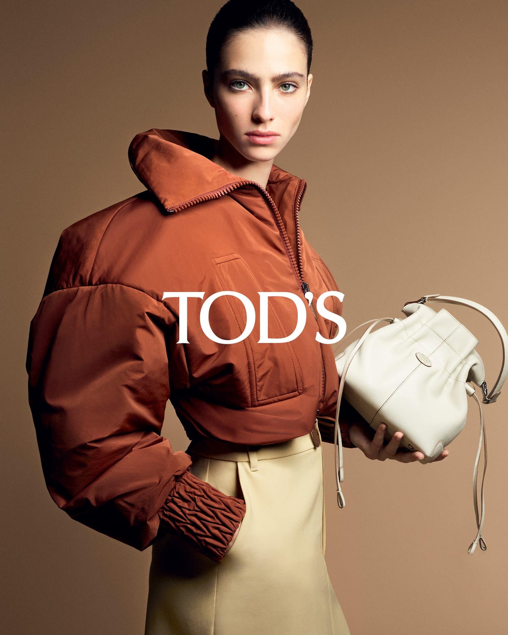 Fashion editorial shot of a woman in a brown jacket and beige skirt, holding a white bucket bag, against a neutral backdrop.