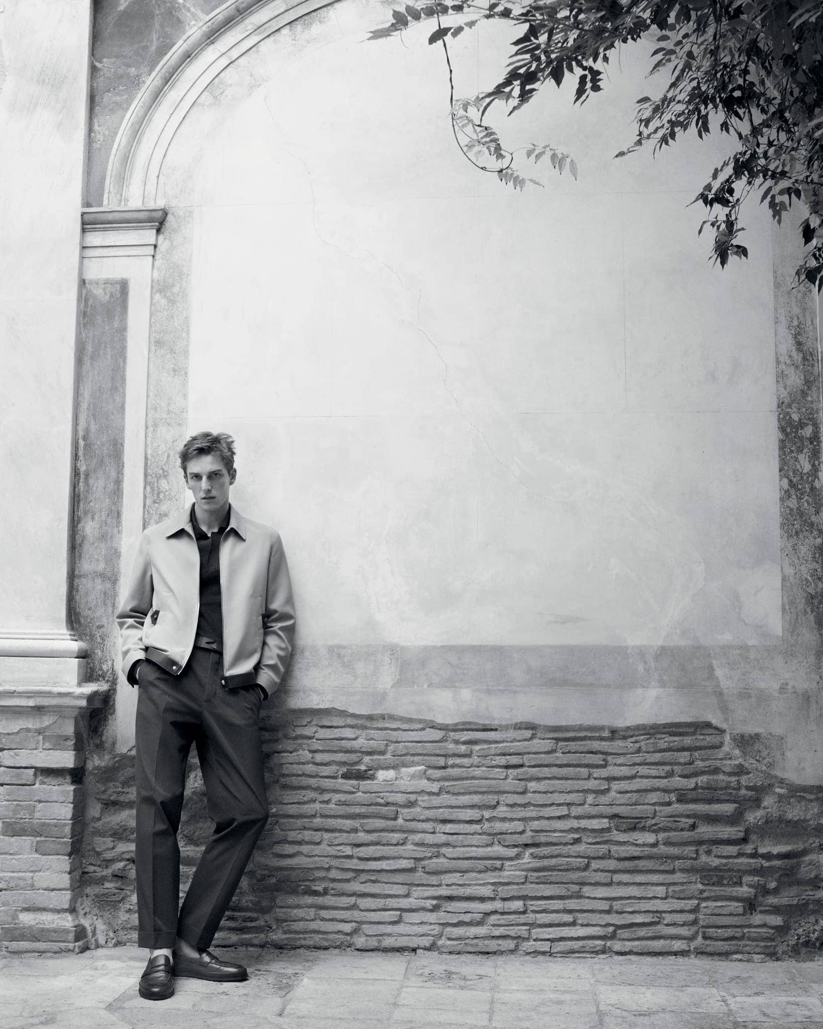 Portrait of a young adult male fashion model leaning against a brick and stucco wall in a beige jacket and black pants outdoors.
